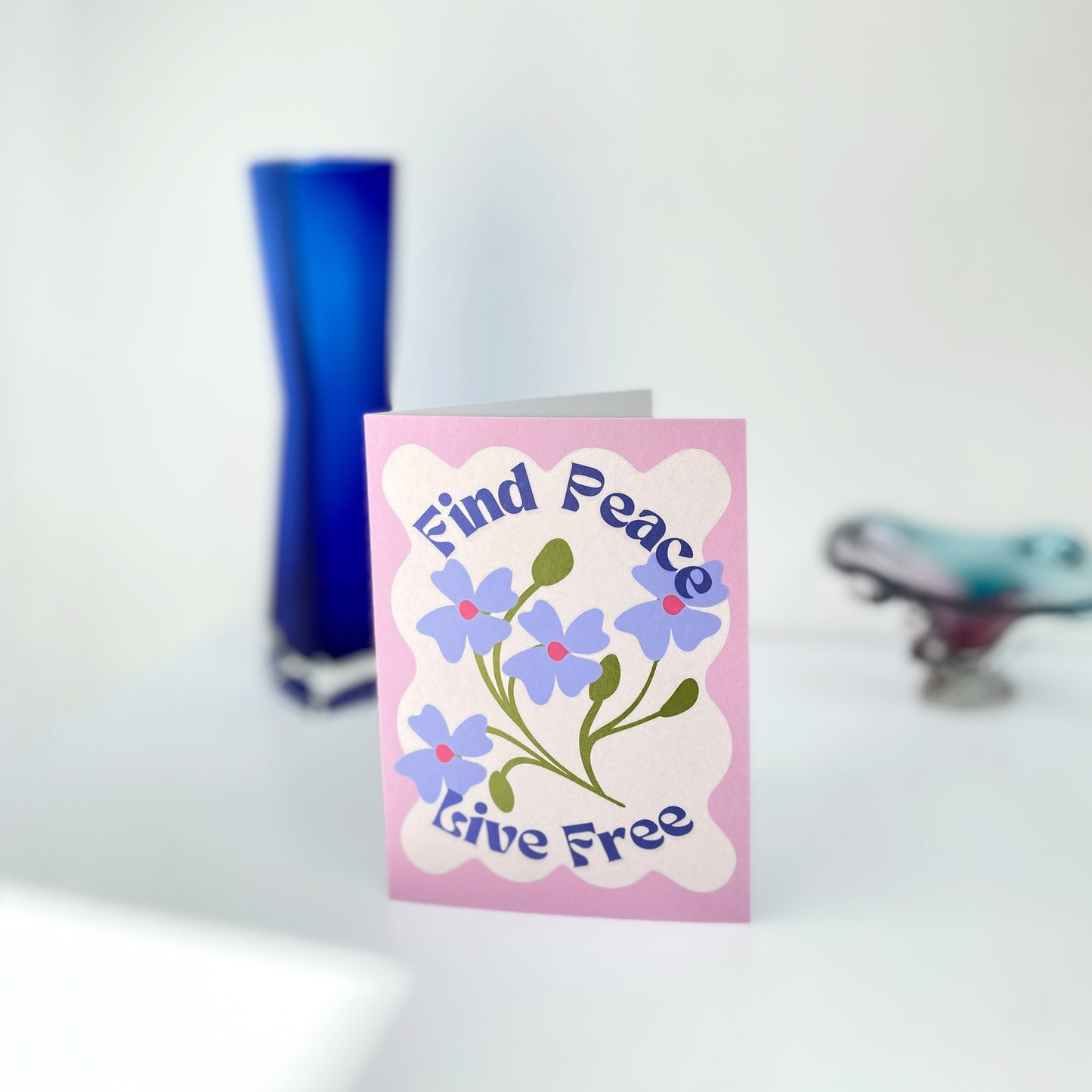 Greeting card with floral design and text 'Find Peace, Live Free' on a pink background, placed on a white surface with a blue vase in the background.