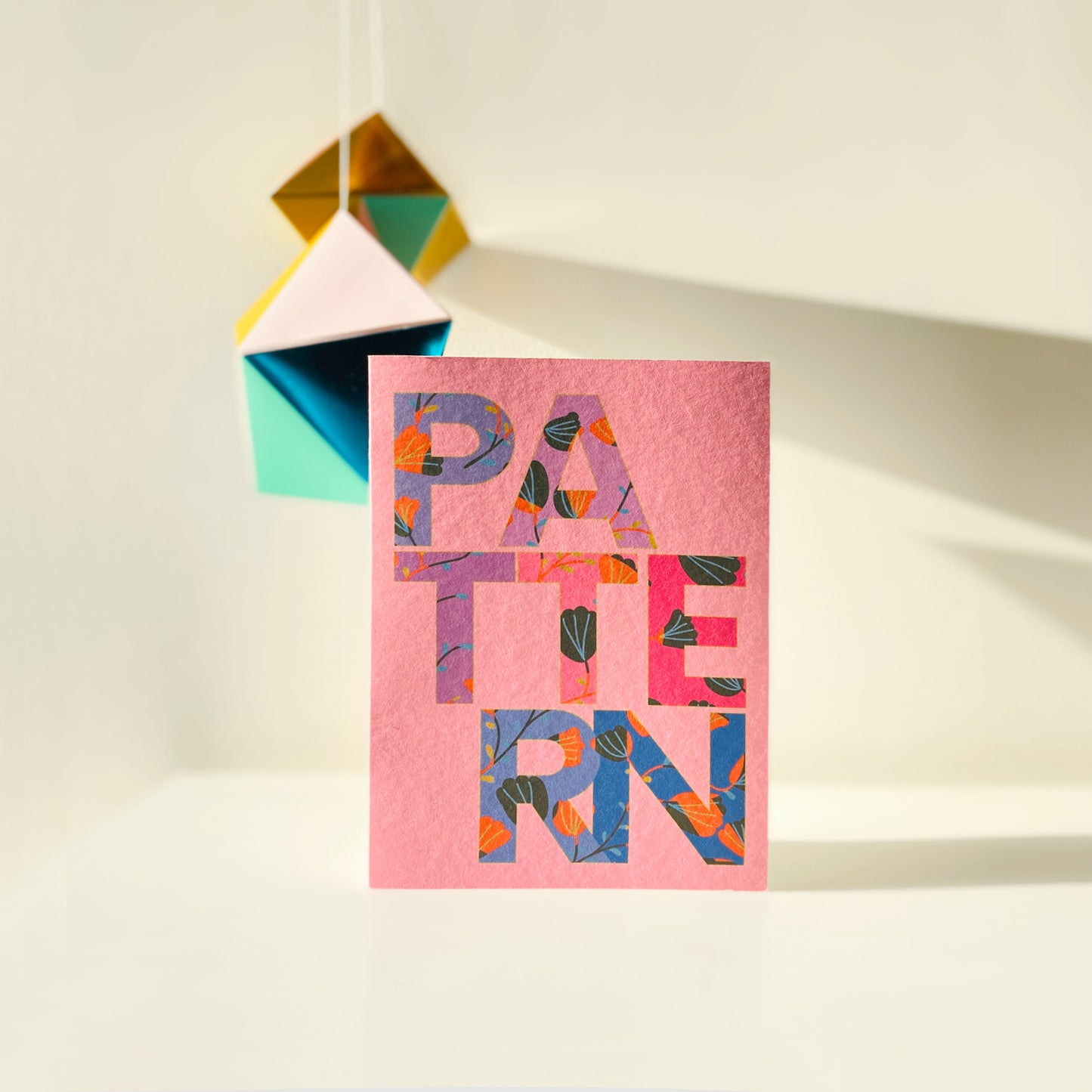 Colorful card with 'PATTERN' text on a pink background, placed on a white surface with a geometric decoration.