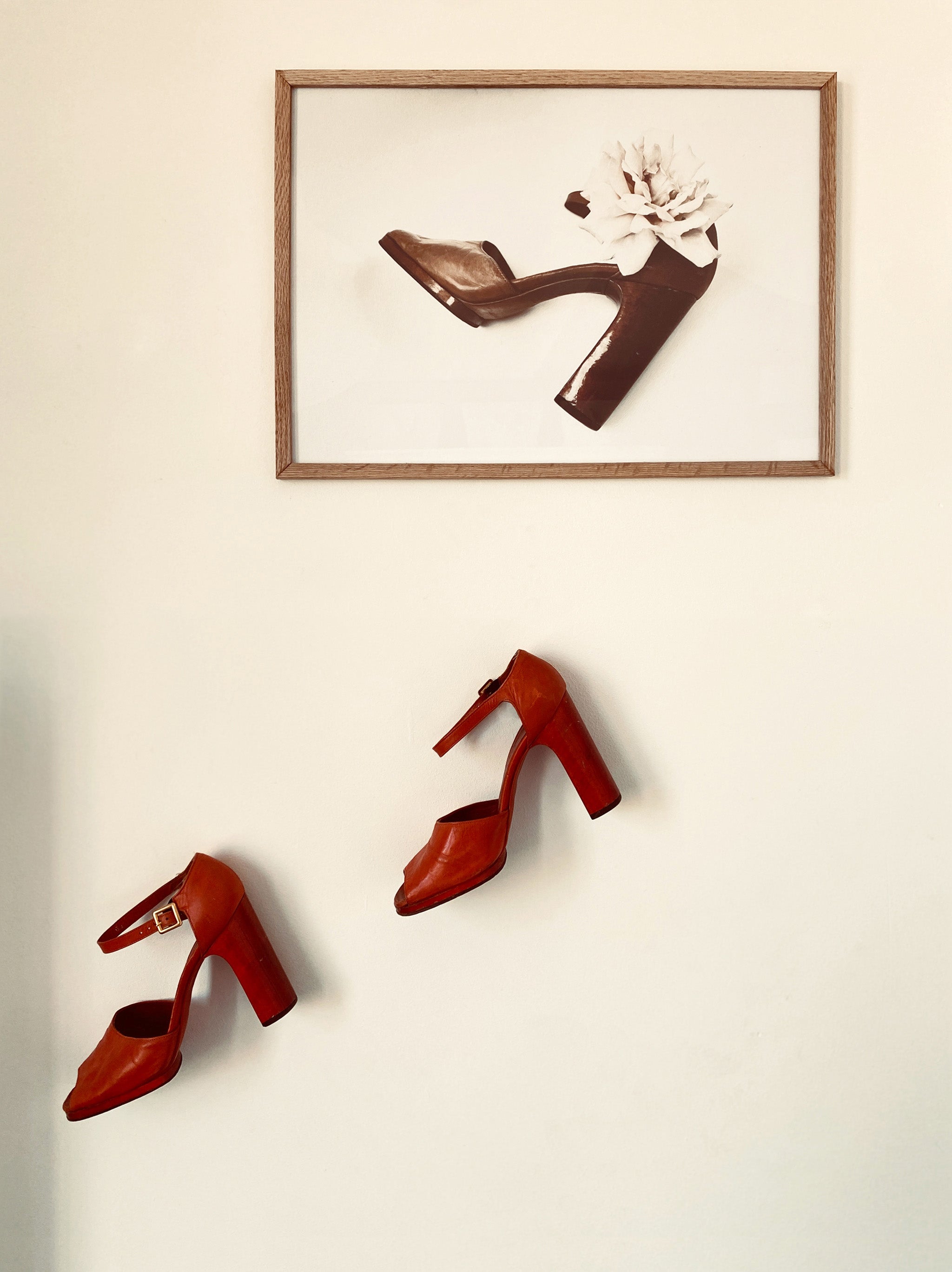Red high-heeled shoes on a white wall with a framed picture of a brown shoe and white flower above.