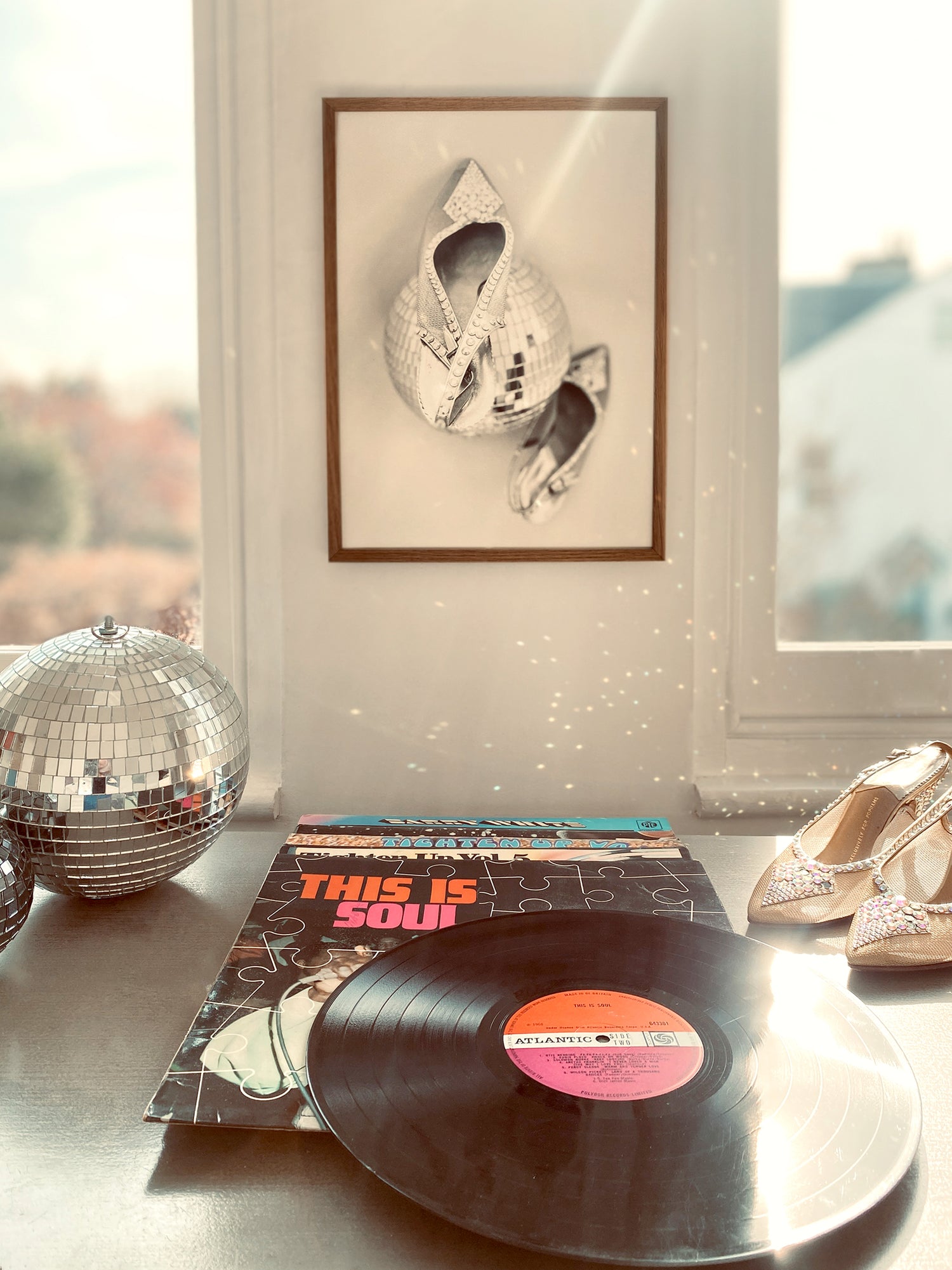 Frame print of shoes and disco ball on wall above Vinyl record with a cover titled 'This is Soul' on a surface with disco ball and shoes.