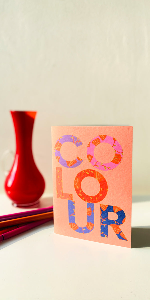 Colorful card with 'COLOUR' text on a light surface with a red vase and pencils.