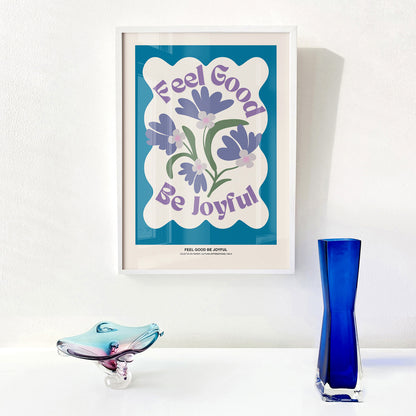 Framed artwork with floral design and "Feel Good Be Joyful" text on a white wall with decorative objects.