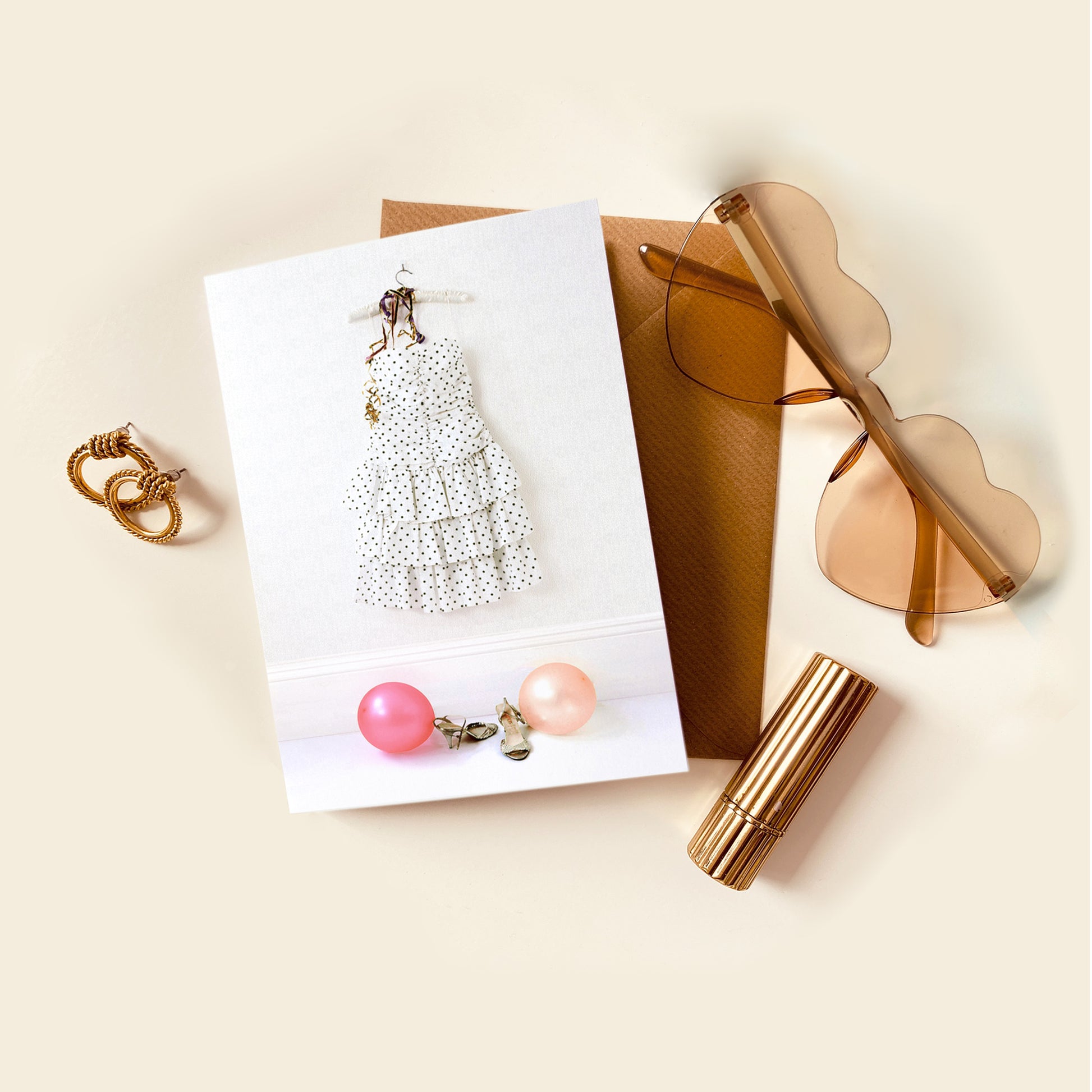 Card with polka dot dress design placed next to earrings, sunglasses and lip gloss on a beige surface