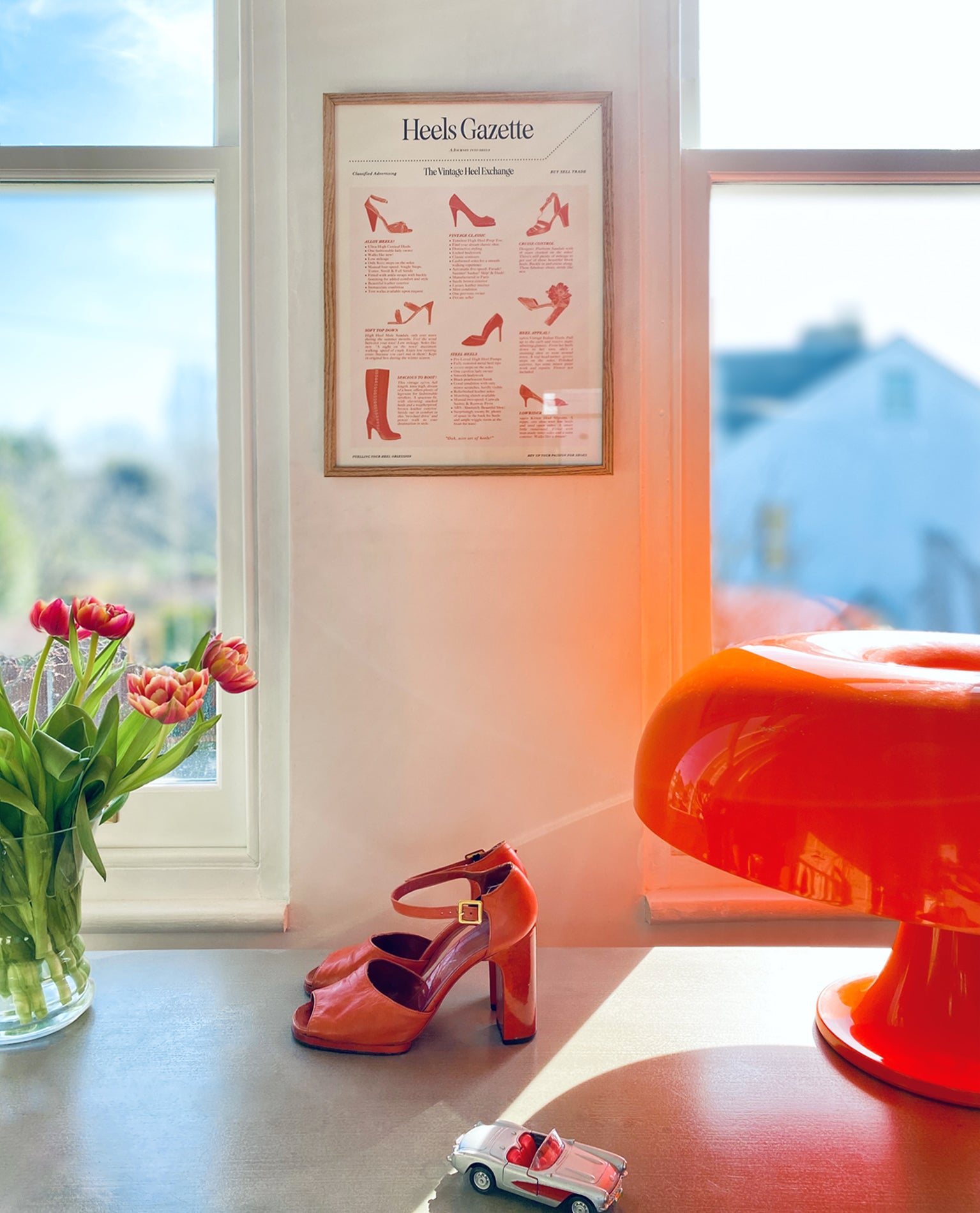 Orange high-heeled shoes on a table with a vase of tulips and a toy car, retro table lamp and framed poster on the wall.