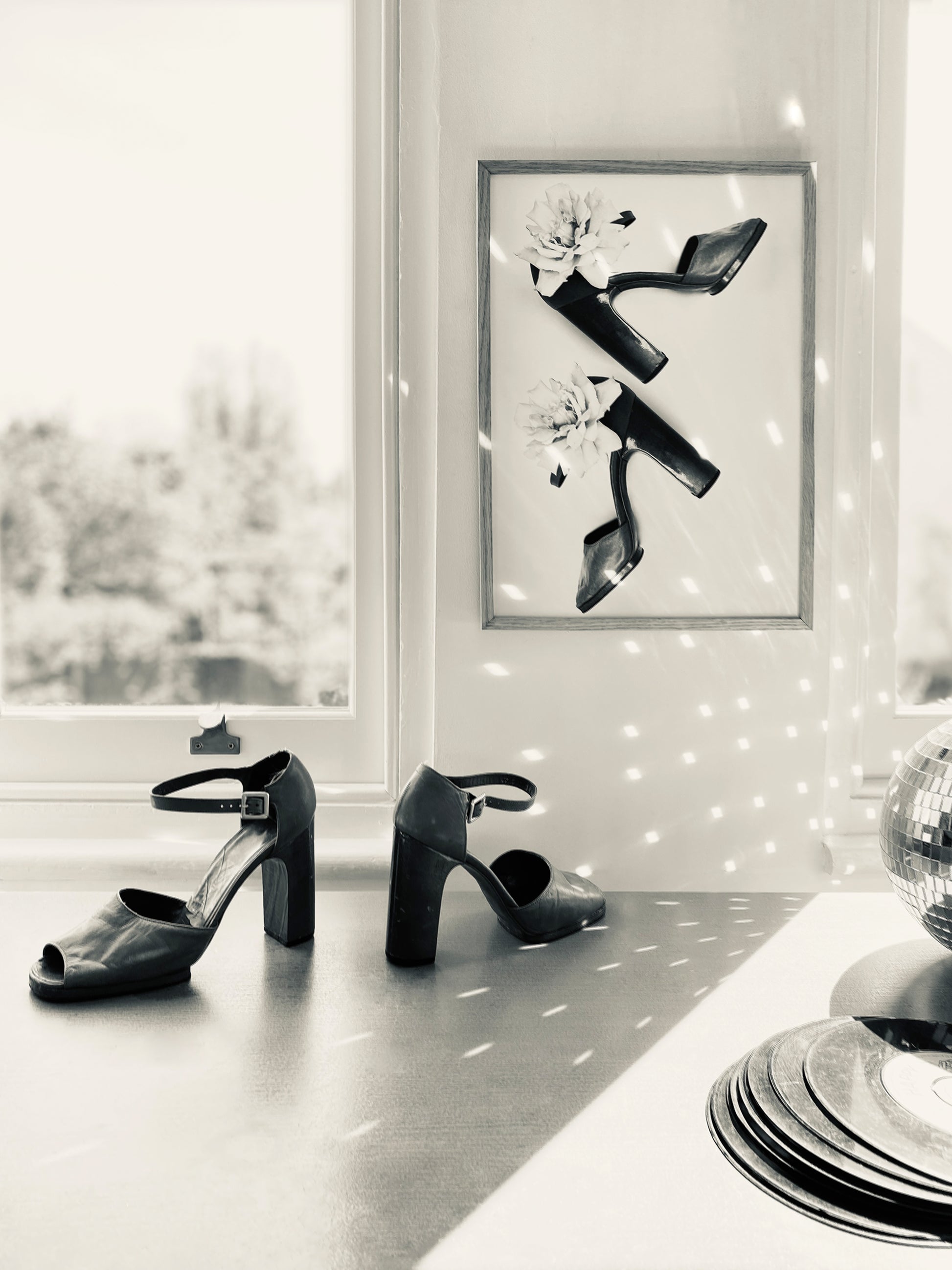 Framed black and white art piece of vintage shoes, vinyl record, disco ball reflecting the light, and matching pair of vintage heels on a surface.