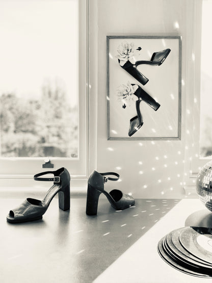 Framed black and white art piece of vintage shoes, vinyl record, disco ball reflecting the light, and matching pair of vintage heels on a surface.
