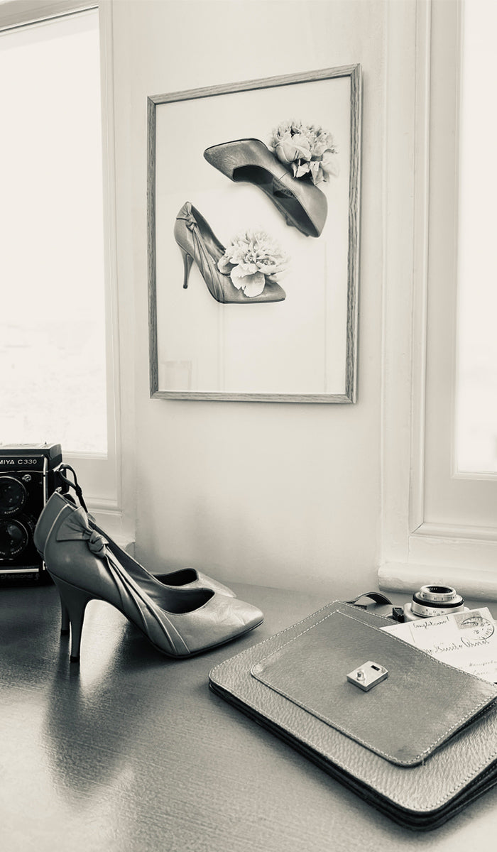 High-heeled shoes on a table with a framed picture of shoes and flowers on the wall.