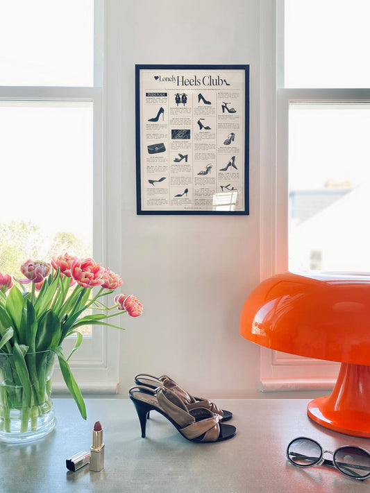 Framed shoe poster on a wall with shoes, lipstick, and sunglasses on a table below.