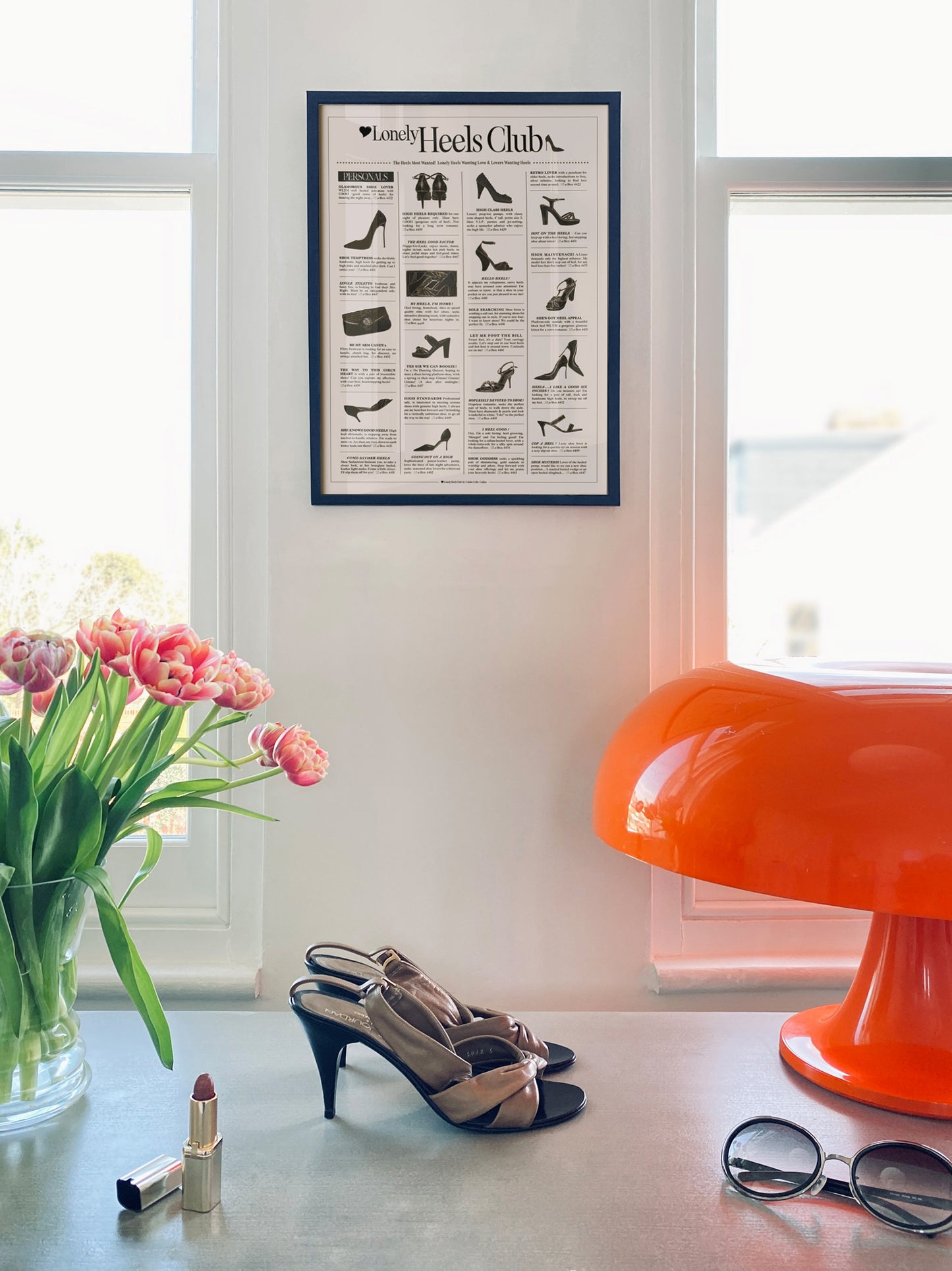 Framed 'Lonely' Heels Club' poster on a wall with high heels, lipstick, and sunglasses on a table.