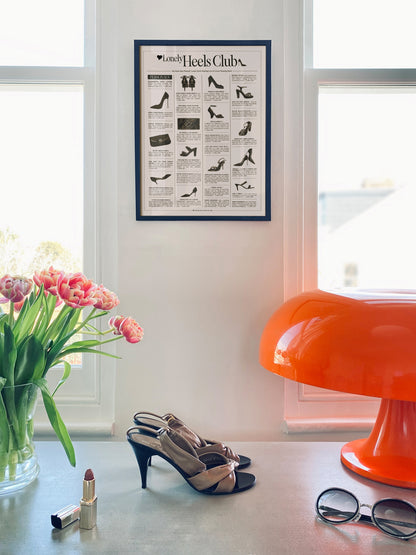 Framed 'Lonely' Heels Club' poster on a wall with high heels, lipstick, and sunglasses on a table.