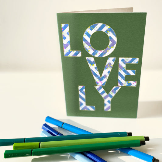 Green card with 'LOVELY' text and pens on a white background