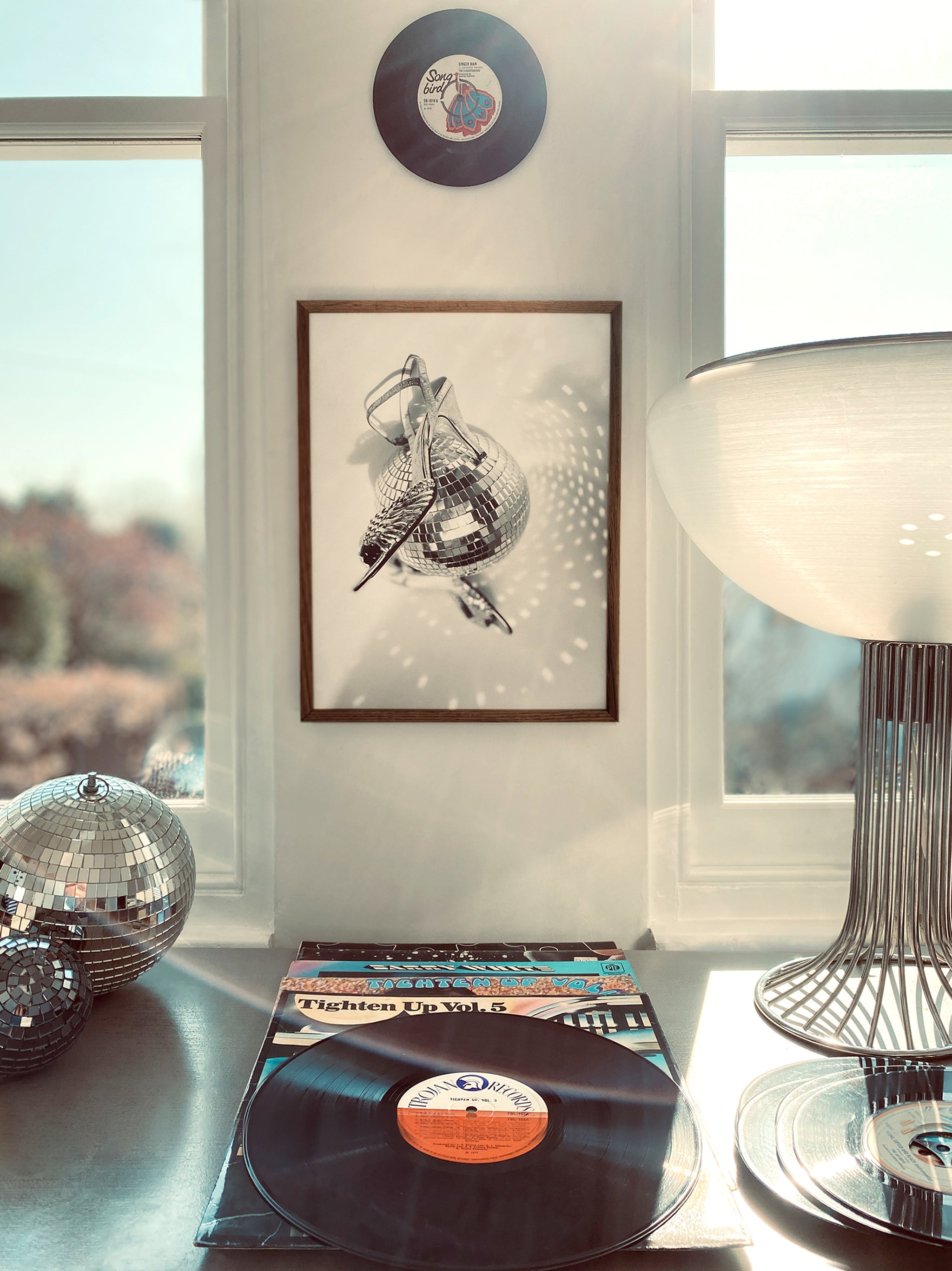 Decorative items including vinyl records, a disco ball, and a lamp on a surface with  framed art of shoes spinning around a disco ball on the wall and window in the background.