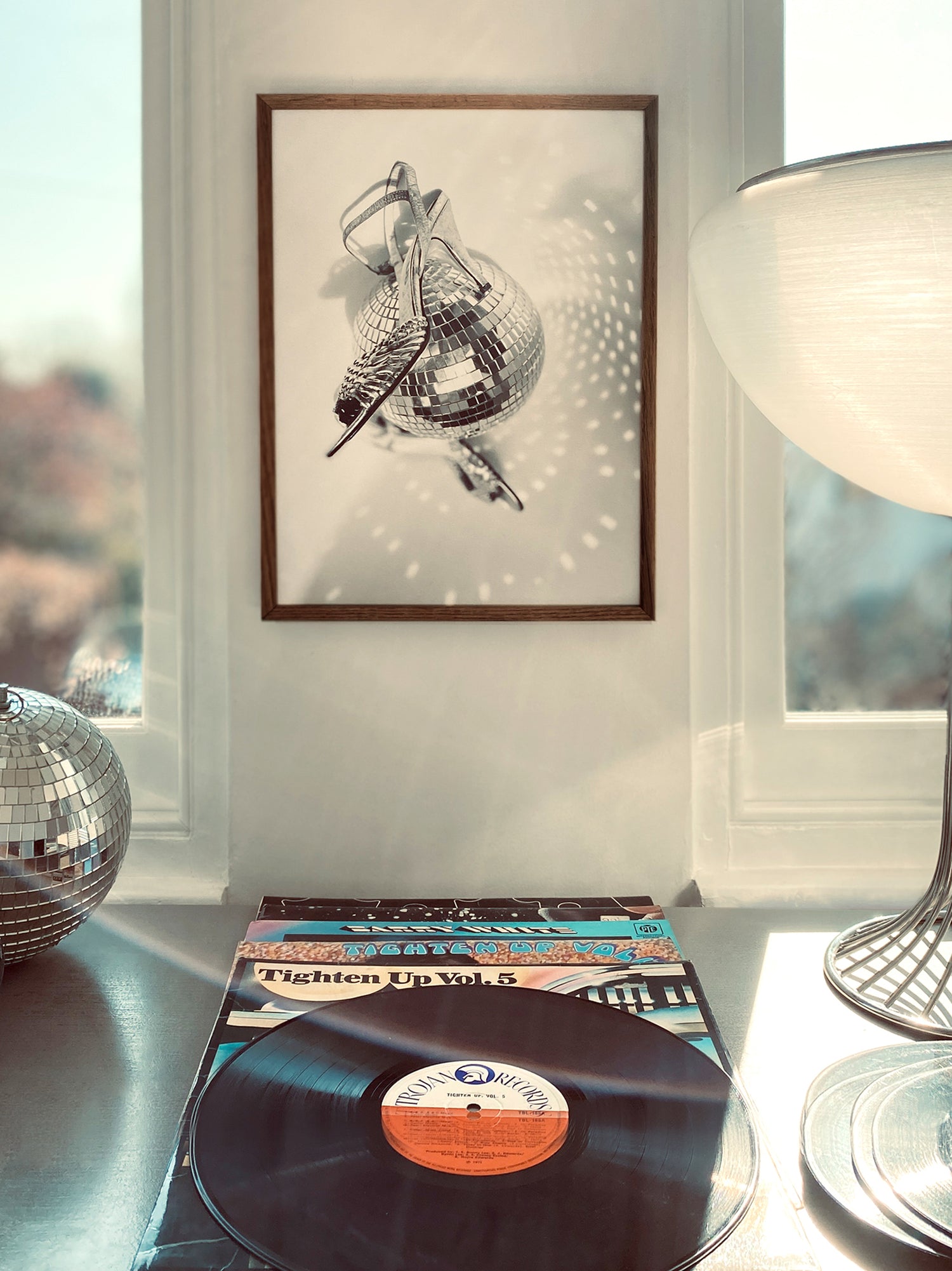 Vinyl record with cover on a surface and framed art of shoes spinning around a disco ball in the background.