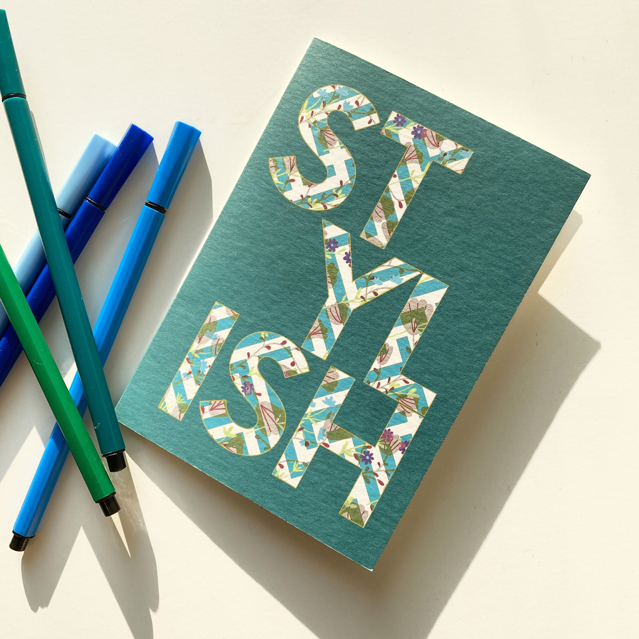 Green greeting card with mosaic letter design next to blue pens on a light background