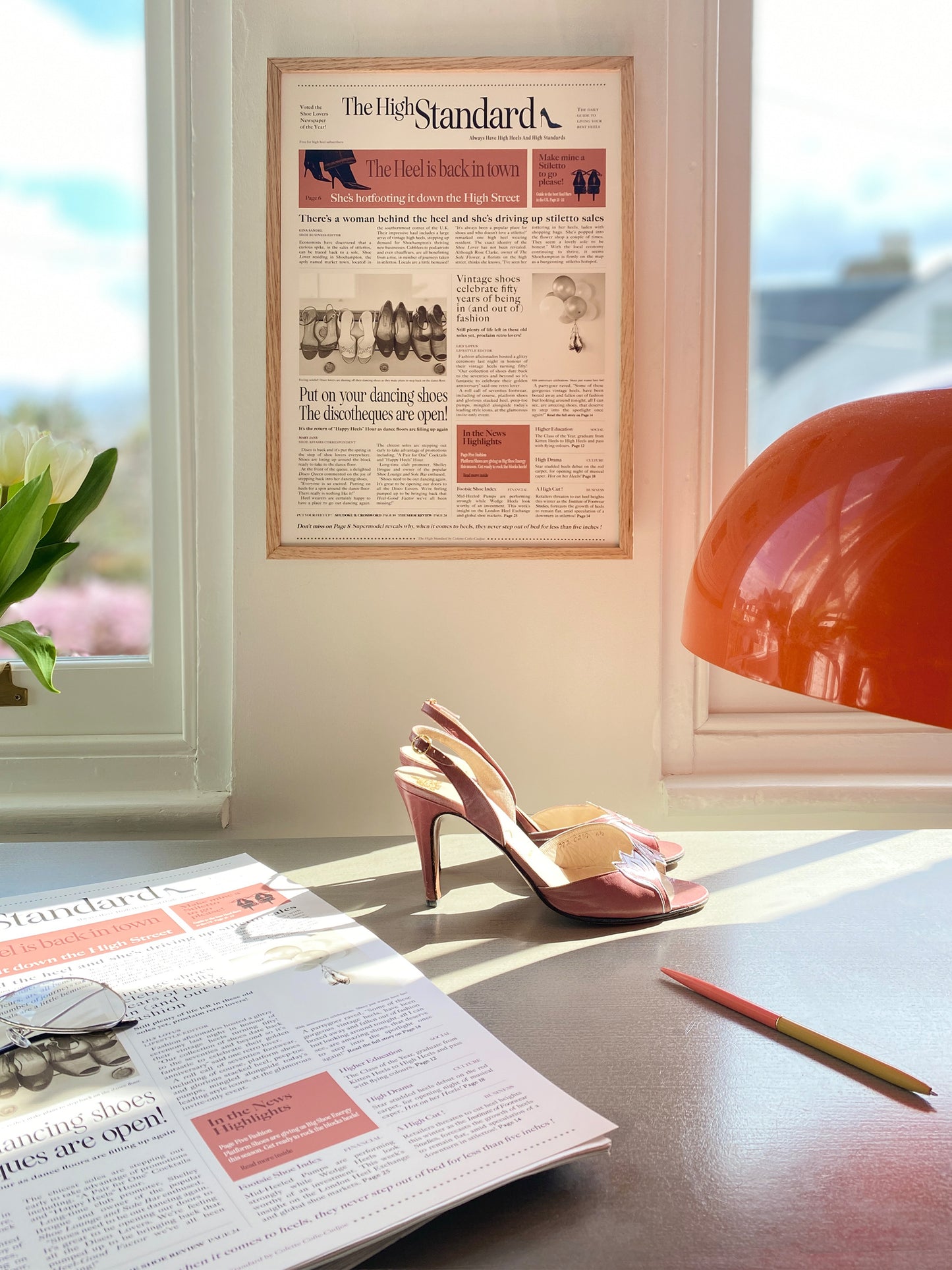 A pair of high heels placed on a table next to a pen and a framed newspaper headline that reads 'The High Standard' featuring articles and stories about shoes.
