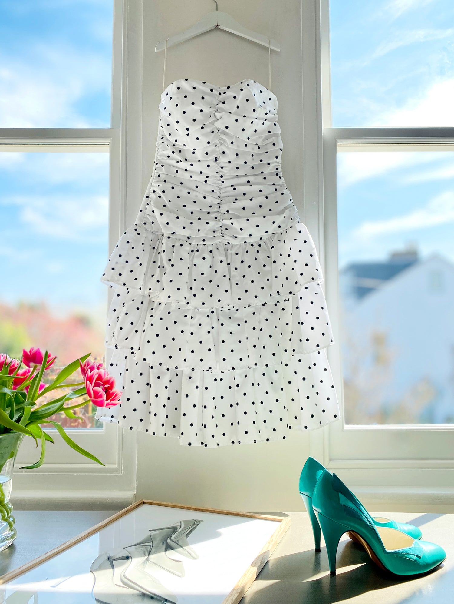 White dress with black polka dots hanging by a window with a pair of teal shoes and flowers on a table.