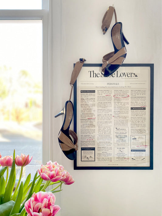 A wall decor featuring a framed print titled 'The Shoe Lover', depicting a text-based personal advertisement for shoes, with a pair of high heels hanging from the wall above the frame, set in a bright room with natural light and pink tulips in the foreground.