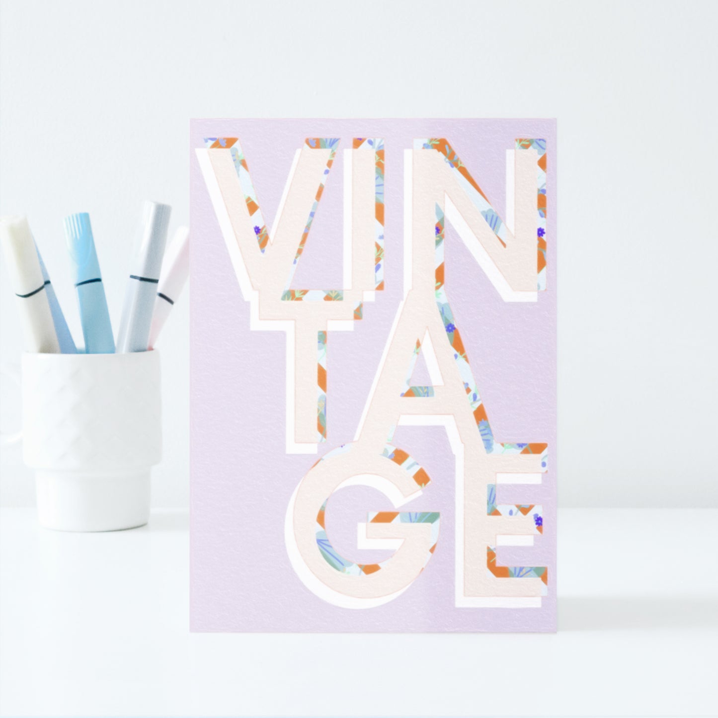 Decorative card with colourful letters spelling VINTAGE on a light purple background, next to a cup with pastel marker pens.