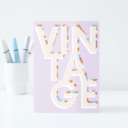 Decorative card with colourful letters spelling VINTAGE on a light purple background, next to a cup with pastel marker pens.